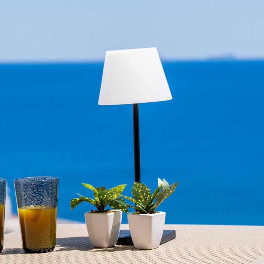 Δύο ποτήρια χυμό πορτοκάλι, δύο μικρά φυτά σε γλάστρες και ένα λευκό επιτραπέζιο φωτιστικό βρίσκονται σε ένα τραπέζι με φόντο το γαλάζιο της θάλασσας και τον ουρανό.