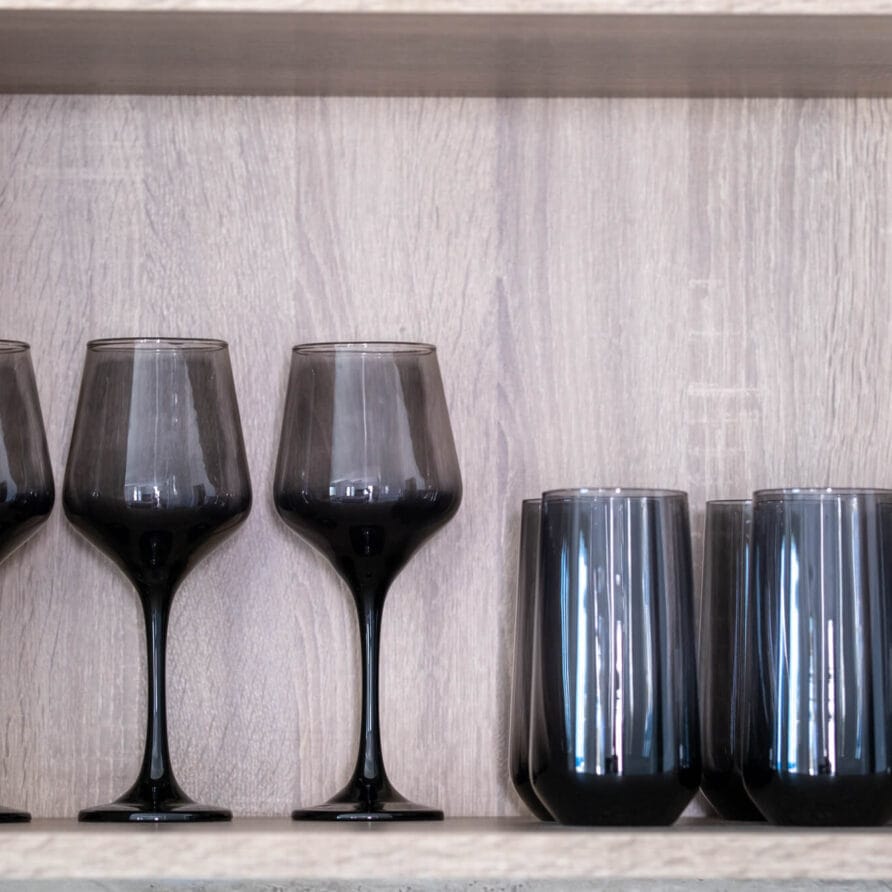 Έξι μαύρα ποτήρια εκτίθενται σε ένα ελαφρύ ξύλινο ράφι — τα τρία είναι ποτήρια κρασιού με στέλεχος και τα τρία είναι πιο κοντά, χωρίς στέλεχος.