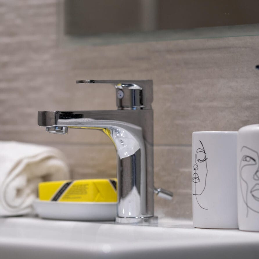 Ένας μοντέρνος νιπτήρας μπάνιου με χρωμιωμένη βρύση, μια διπλωμένη λευκή πετσέτα, μια σαπουνοθήκη, ένα φλιτζάνι και ένας διανομέας σαπουνιού, και τα δύο με μινιμαλιστικά σχέδια γραμμικής τέχνης προσώπου.