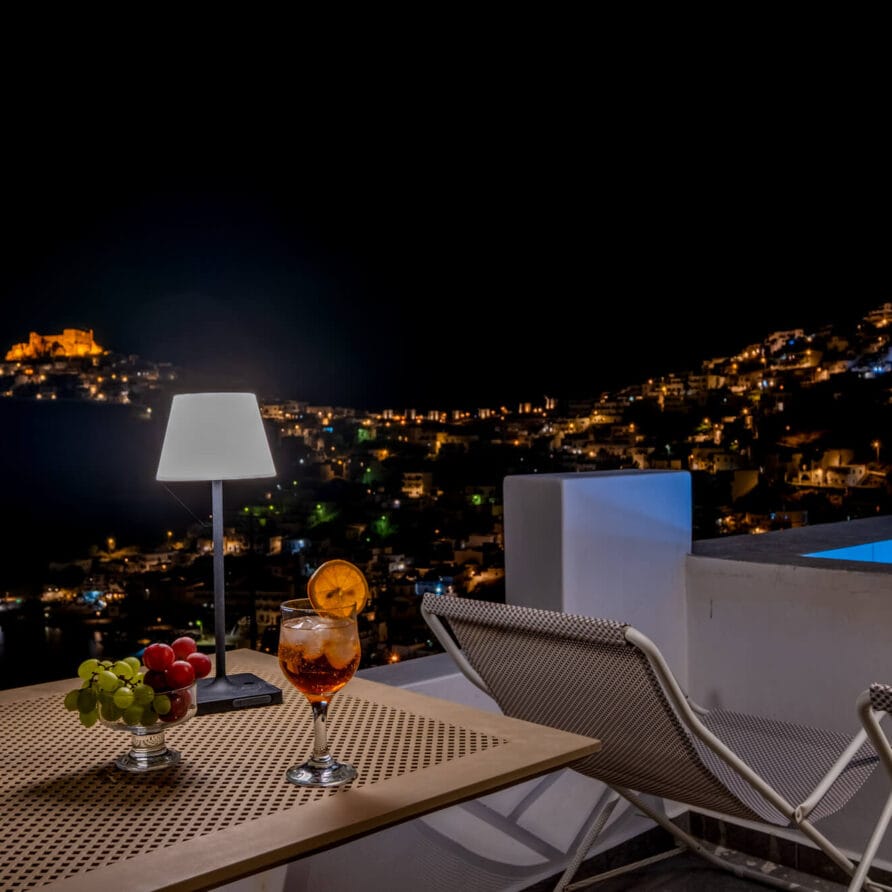 Νυχτερινή θέα μιας πόλης στην πλαγιά ενός λόφου με φωτισμένα κτίρια, δύο ξαπλώστρες, ένα τραπέζι με ποτά και σταφύλια και ένα μικρό αναμμένο φωτιστικό δίπλα σε μια πισίνα.