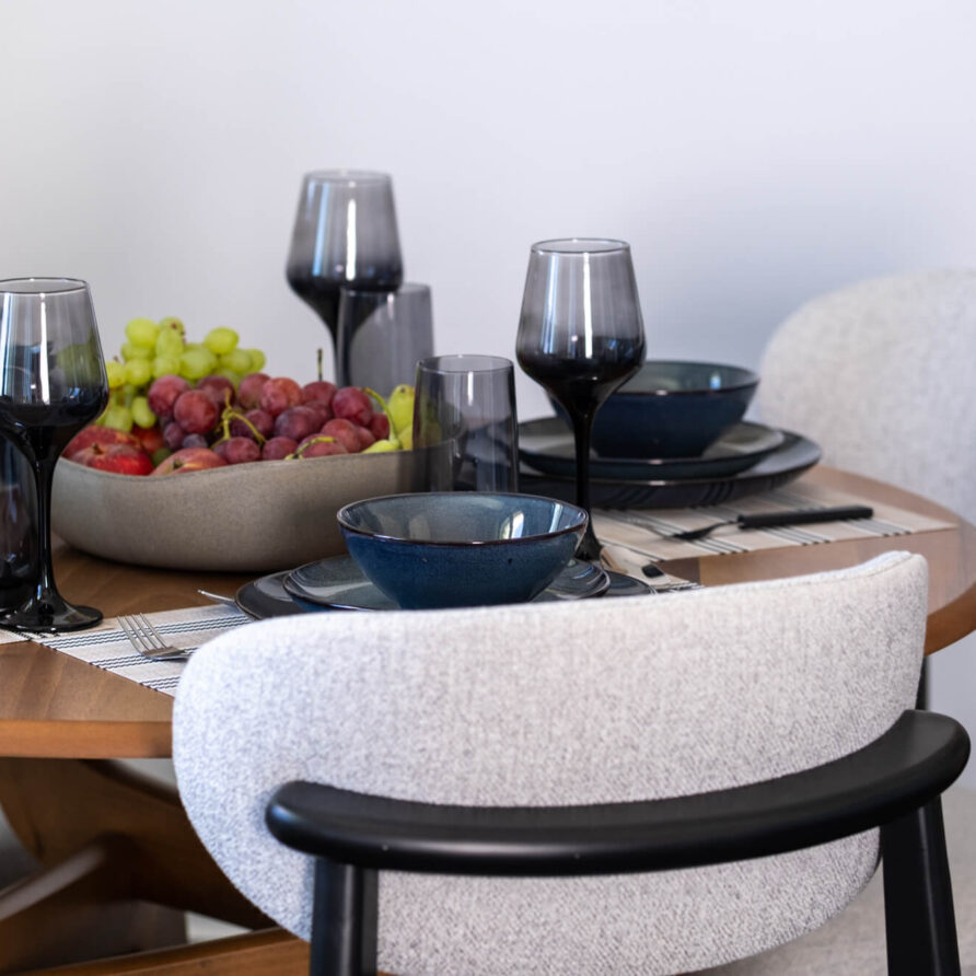 Ένα στρογγυλό ξύλινο τραπέζι φαγητού με μαύρα γυάλινα σκεύη, σκούρα πιάτα και ένα μπολ με κόκκινα και πράσινα σταφύλια, περιτριγυρισμένο από ταπετσαρισμένες καρέκλες.