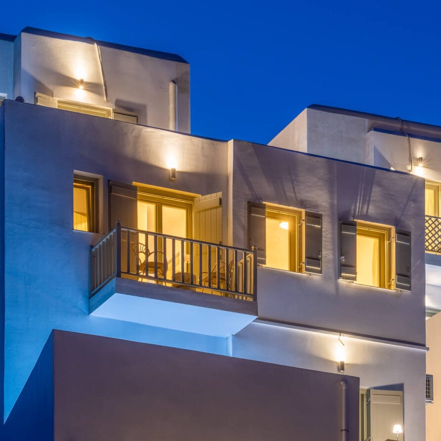 Σύγχρονο λευκό κτίριο με πολλαπλά μπαλκόνια και μεγάλα παράθυρα που φωτίζονται από ζεστά φώτα το σούρουπο με φόντο έναν καθαρό γαλάζιο ουρανό.