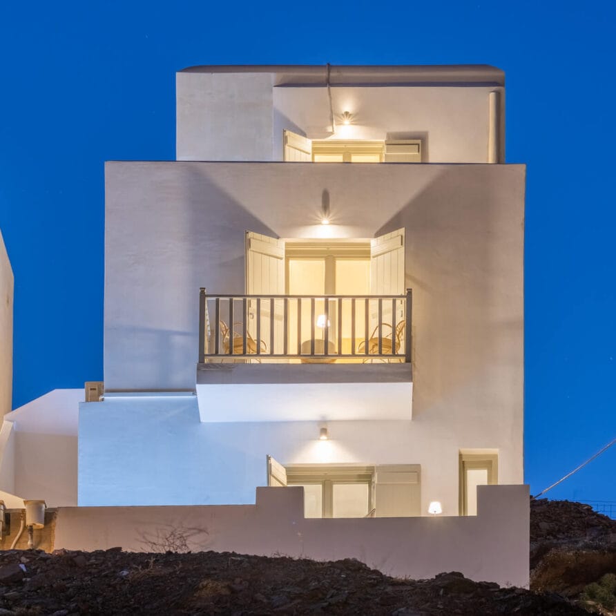 Σύγχρονο λευκό τριώροφο σπίτι με μπαλκόνια και φωτισμένα εξωτερικά φώτα, φωτογραφημένο το σούρουπο με φόντο έναν βαθύ μπλε ουρανό.