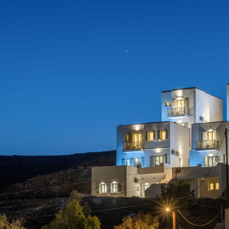 Λευκά, μοντέρνα, πολυώροφα κτίρια με φωτισμένα μπαλκόνια στην πλαγιά ενός λόφου το σούρουπο, με φόντο έναν καθαρό, βαθύ γαλάζιο ουρανό. Δέντρα και ηλεκτροφόρα καλώδια είναι ορατά στο πρώτο πλάνο.