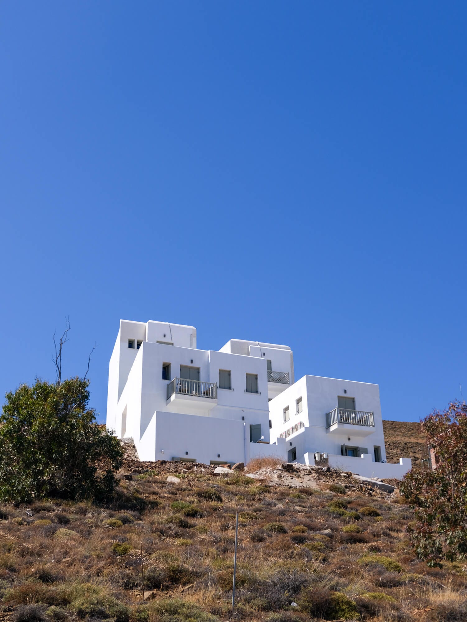 White, cubic buildings with balconies sit on a dry, rocky hillside under a clear blue sky, surrounded by sparse vegetation.
