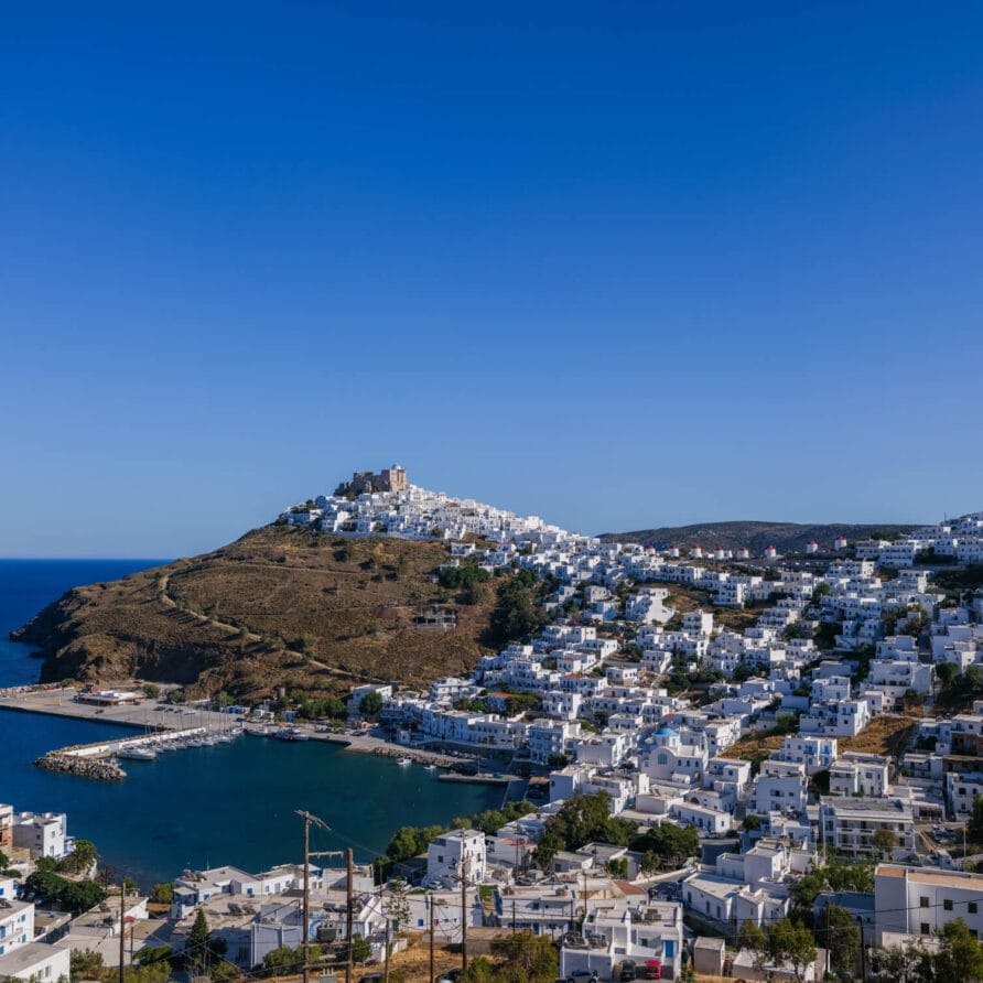 Μια παραθαλάσσια πόλη με λευκά κτίρια στις πλαγιές των λόφων έχει θέα στη θάλασσα κάτω από έναν καταγάλανο ουρανό, με ένα περίοπτο κάστρο στην κορυφή του κεντρικού λόφου.