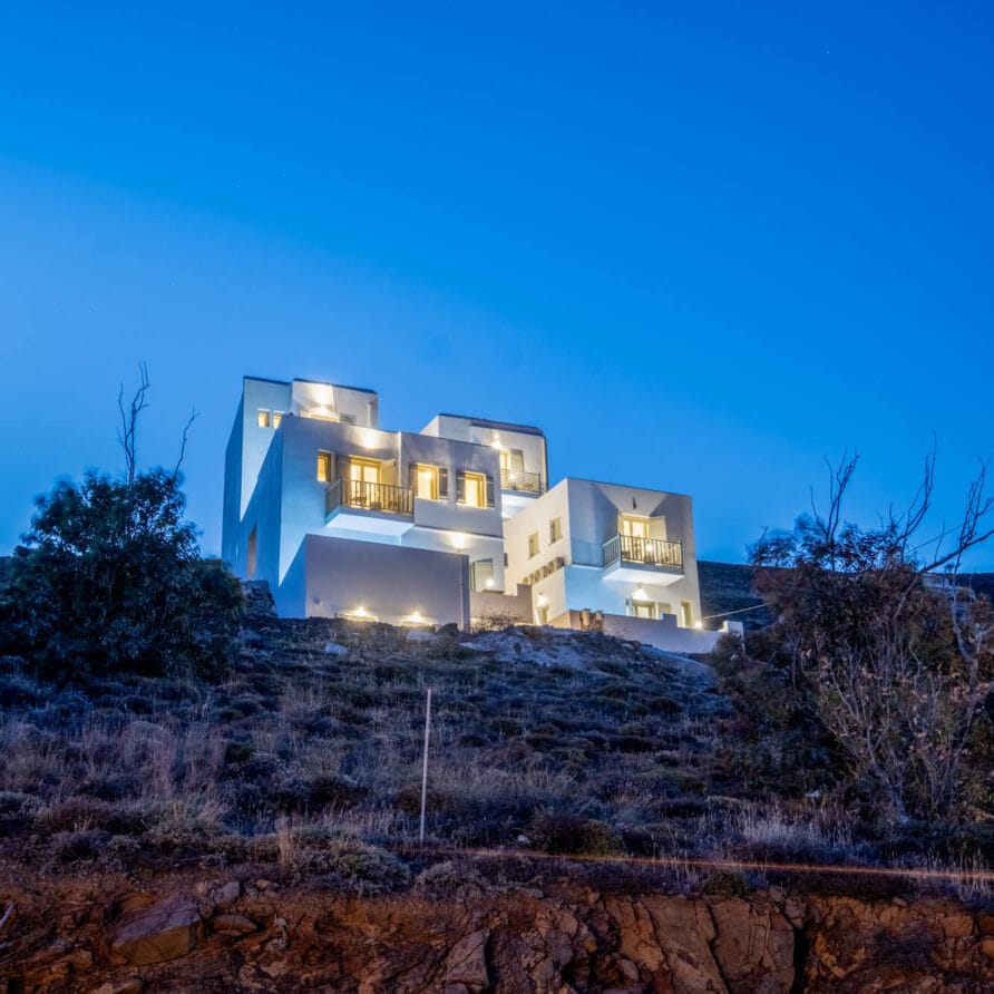 Σύγχρονο λευκό πολυώροφο σπίτι με φωτισμένα παράθυρα βρίσκεται στην πλαγιά ενός λόφου το σούρουπο, περιτριγυρισμένο από αραιή βλάστηση και βραχώδες προσκήνιο.