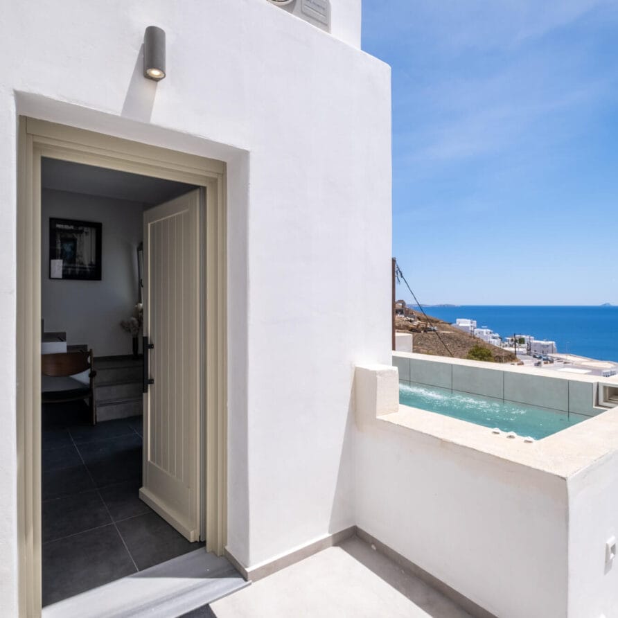 Ένα λευκό κτίριο με μια ανοιχτή πόρτα οδηγεί σε ένα δωμάτιο, δίπλα σε μια μικρή πισίνα σε μια βεράντα με θέα στη θάλασσα και το παράκτιο τοπίο κάτω από έναν καταγάλανο ουρανό.