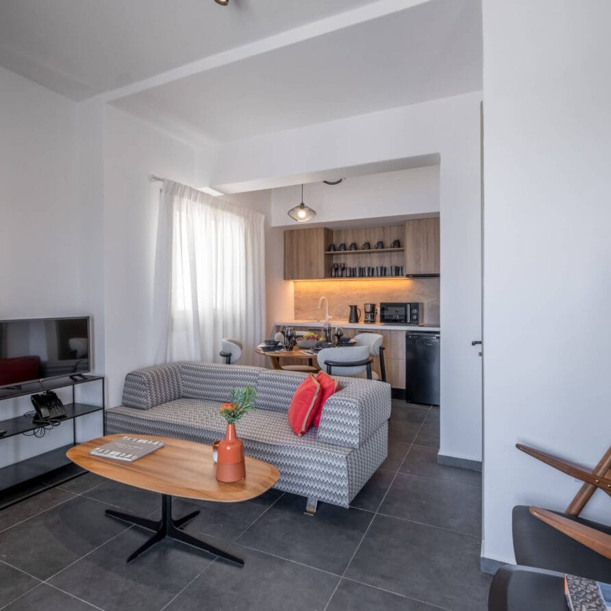 Μοντέρνο σαλόνι με γκρι καναπέ, μικρό τραπεζάκι σαλονιού, τηλεόραση και ανοιχτή κουζίνα. Φως μπαίνει από ένα παράθυρο και ξύλινες καρέκλες βρίσκονται δίπλα σε μια σκάλα.