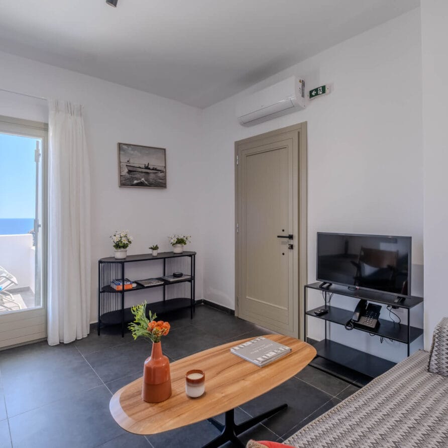 Μοντέρνο σαλόνι με καναπέ, τραπεζάκι σαλονιού, τηλεόραση και ράφια, με μεγάλα παράθυρα και πόρτα που ανοίγει σε μπαλκόνι με θέα στη θάλασσα.