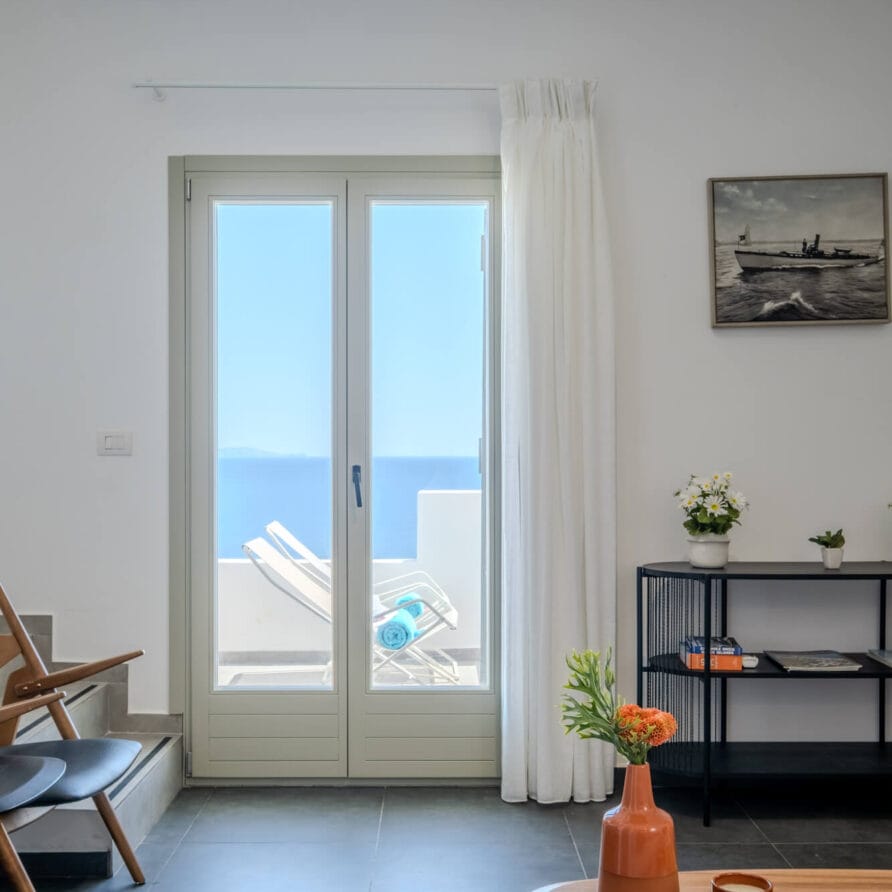 Ένα φωτεινό δωμάτιο με γυάλινη πόρτα που οδηγεί σε μια βεράντα με θέα στη θάλασσα, επιπλωμένο με καρέκλες, ένα μικρό τραπέζι και διακοσμητικά αντικείμενα.