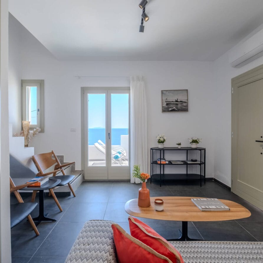 Μοντέρνο σαλόνι με μινιμαλιστικά έπιπλα, ένα μικρό τραπέζι και μια επιτοίχια τηλεόραση, με πόρτα και παράθυρο που ανοίγουν σε μπαλκόνι με θέα στη θάλασσα στο βάθος.