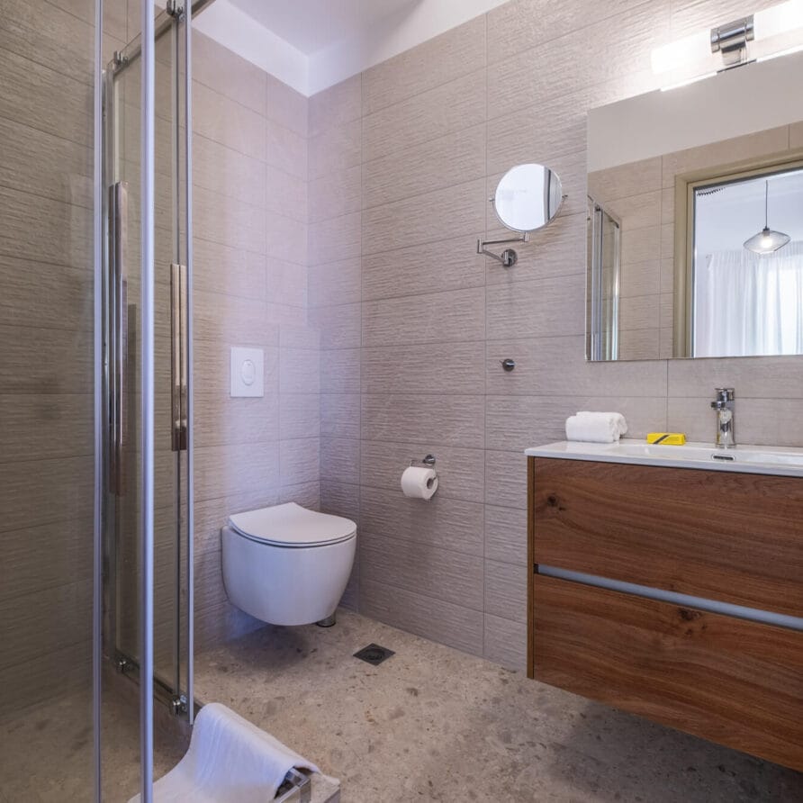 Μοντέρνο μπάνιο με γυάλινη ντουζιέρα, επιτοίχια τουαλέτα, ξύλινο νιπτήρα, μεγάλο καθρέφτη, πετσέτα στο πάτωμα και πλακάκια σε ουδέτερους τόνους.