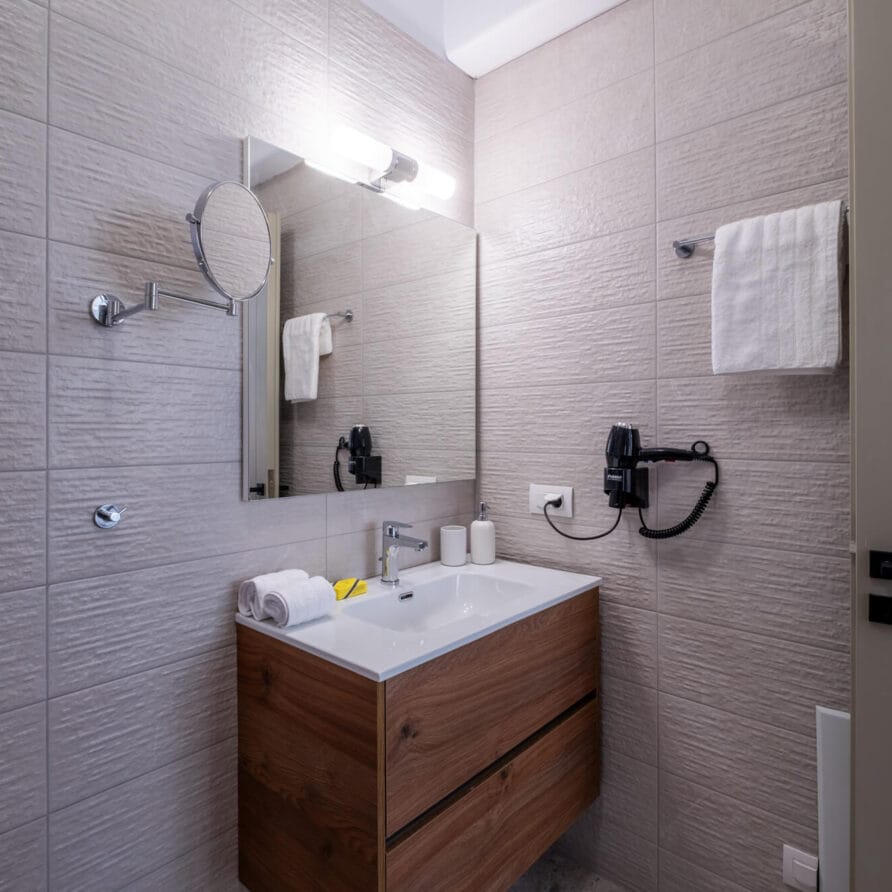 Μοντέρνο μπάνιο με ανοιχτόχρωμους γκρι τοίχους με πλακάκια, ξύλινο νιπτήρα με λευκό νιπτήρα, επιτοίχιο καθρέφτη, τοποθετημένο πιστολάκι μαλλιών, κρεμάστρες για πετσέτες και κλειστή πόρτα.