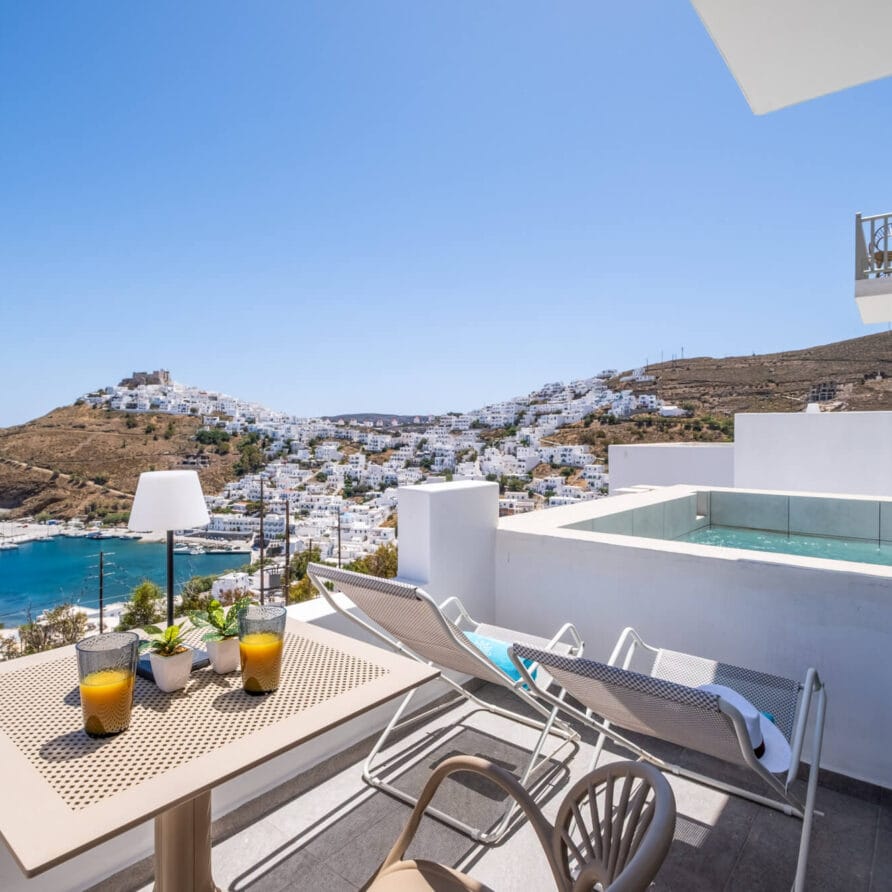 Ένα μπαλκόνι με τραπέζι, καρέκλες και ποτά έχει θέα σε μια παραθαλάσσια πόλη με λευκά κτίρια, μια πλαγιά λόφου και τη θάλασσα. Στο μπαλκόνι υπάρχει μια μικρή πισίνα.