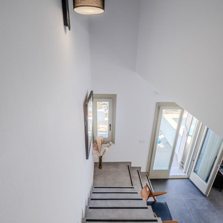 Θέα από την κορυφή μιας εσωτερικής σκάλας με γκρίζα σκαλοπάτια, ένα επιτοίχιο φωτιστικό, ένα βάζο σε ένα μικρό τραπέζι και το φως του ήλιου που μπαίνει μέσα από ανοιχτές γυάλινες πόρτες που οδηγούν έξω.