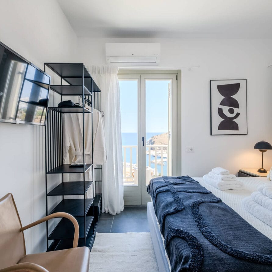 Μοντέρνο δωμάτιο ξενοδοχείου με κρεβάτι, έργα τέχνης τοίχου, τηλεόραση, ανοιχτή ντουλάπα και μπαλκονόπορτα που προσφέρει θέα στη θάλασσα και την ακτογραμμή έξω.