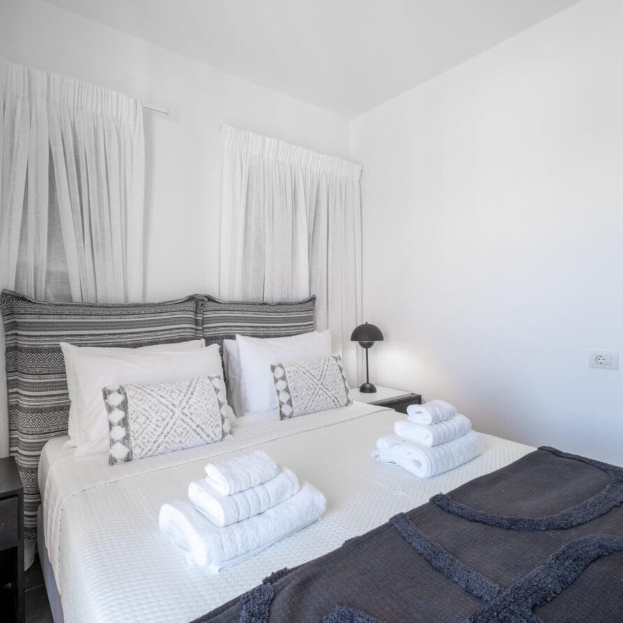 Μινιμαλιστικό υπνοδωμάτιο με διπλό κρεβάτι, ριγέ κεφαλάρι, δύο κομοδίνα με μαύρα φωτιστικά, λευκές κουρτίνες, διπλωμένες πετσέτες στο κρεβάτι και θέα σε ένα διπλανό δωμάτιο.
