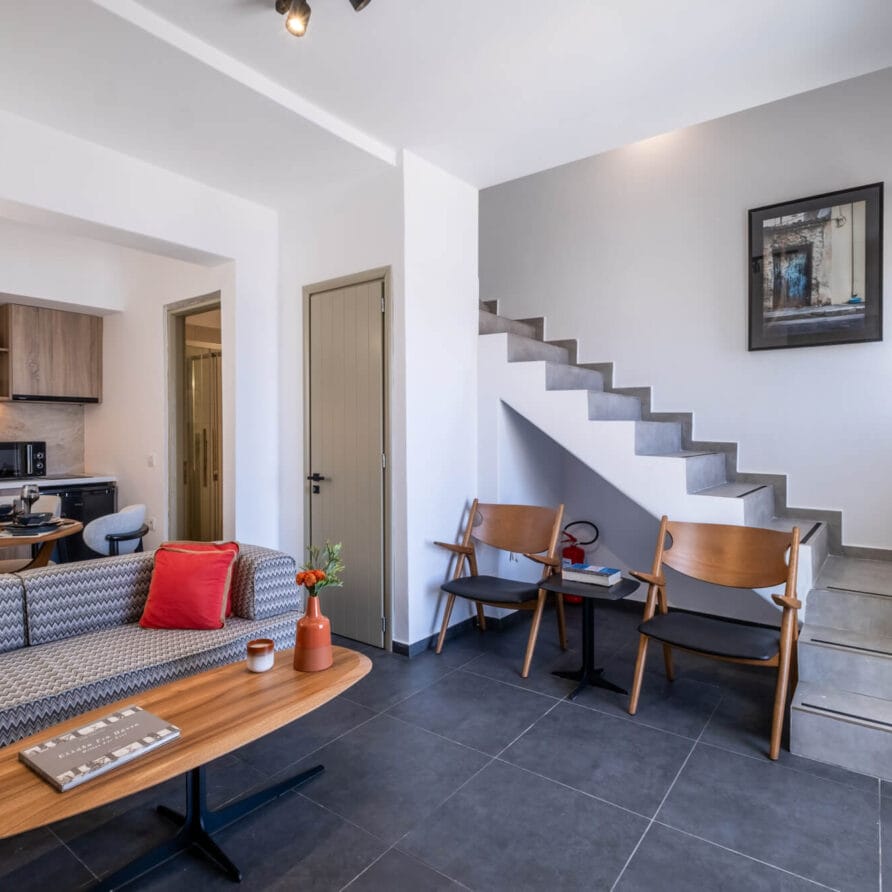 Μοντέρνο σαλόνι με γκρι καναπέ, ξύλινο τραπεζάκι σαλονιού, μικρή κουζίνα, δύο καρέκλες δίπλα στη σκάλα και έργα τέχνης τοίχου πάνω από τις σκάλες.