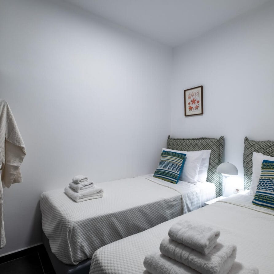 Ένα όμορφα διαμορφωμένο υπνοδωμάτιο με δύο μονά κρεβάτια, ριγέ μαξιλάρια, διπλωμένες πετσέτες, μια μπουρνούζι κρεμασμένη σε ένα ράφι και δύο πλαισιωμένα έργα τέχνης σε έναν λευκό τοίχο.