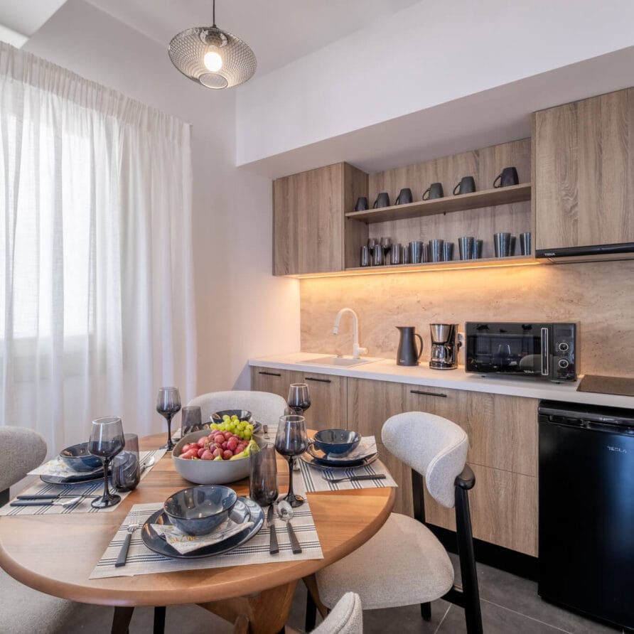 Μοντέρνα κουζίνα με μικρό σετ τραπεζαρίας για τέσσερα άτομα, ξύλινα ντουλάπια, συσκευές στον πάγκο και παράθυρο με διαφανείς κουρτίνες που επιτρέπουν την είσοδο φυσικού φωτός.
