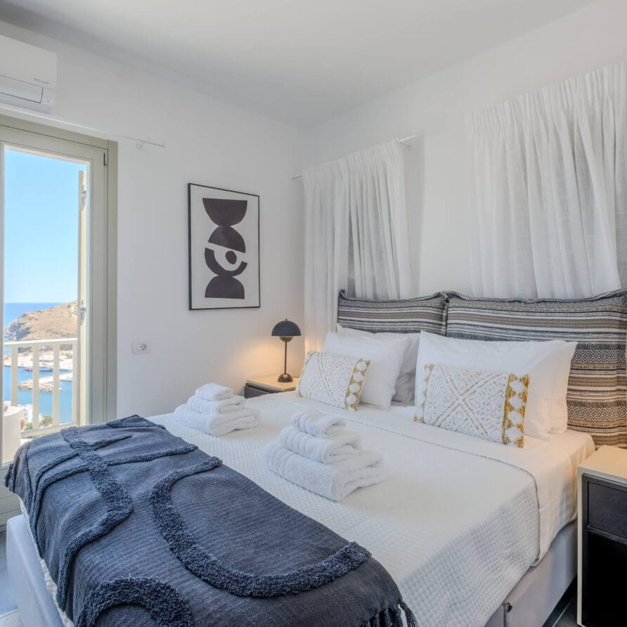 Φωτεινό υπνοδωμάτιο με διπλό κρεβάτι, διακοσμητικά μαξιλάρια, δύο μαύρα φωτιστικά κομοδίνου, διπλωμένες πετσέτες και μπαλκονόπορτα που προσφέρει θέα στη θάλασσα και την ακτογραμμή.