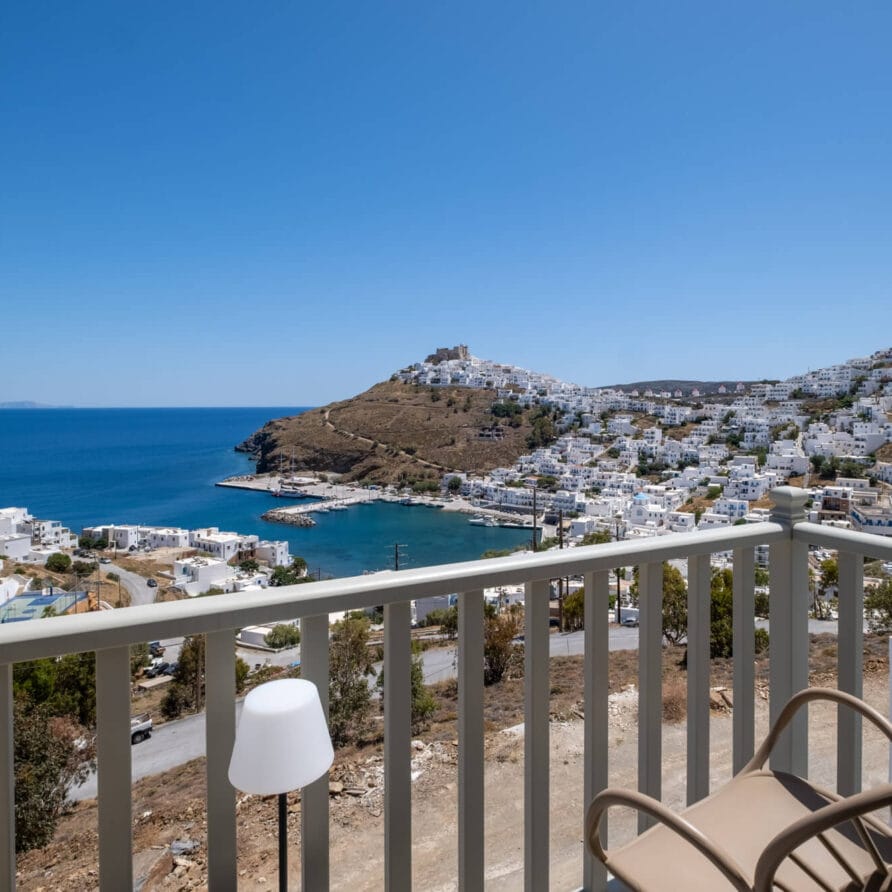 Θέα από μπαλκόνι με θέα σε μια παραθαλάσσια πόλη με λευκά κτίρια στην πλαγιά ενός λόφου, γαλάζια θάλασσα και καθαρό ουρανό. Δύο καρέκλες και μια λάμπα είναι ορατές στο πρώτο πλάνο.