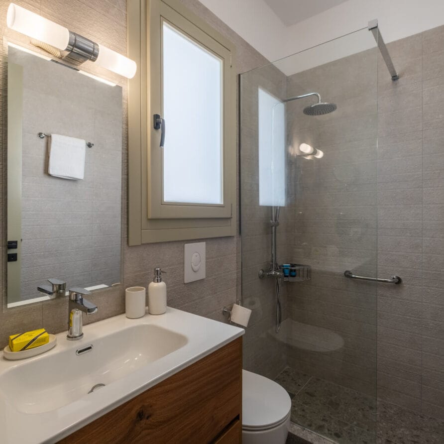 Μοντέρνο μπάνιο με νιπτήρα, καθρέφτη, επιτοίχιο φωτιστικό, αδιαφανές παράθυρο, γυάλινη καμπίνα ντους, πετσέτα και είδη προσωπικής υγιεινής στον πάγκο.