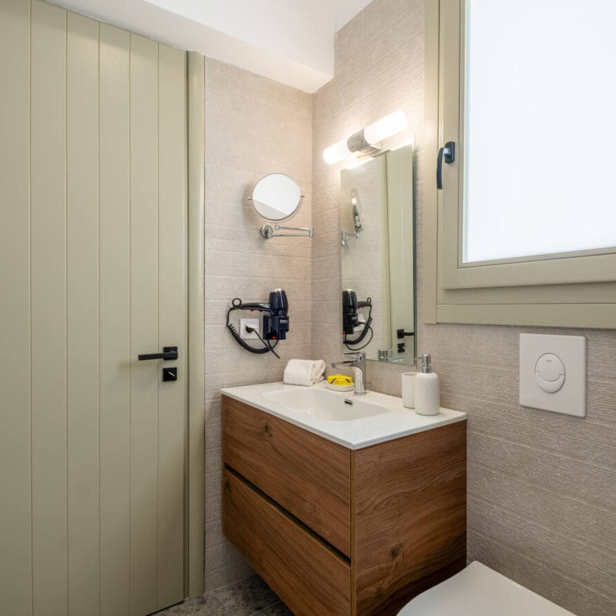 Μοντέρνο μπάνιο με ξύλινο νιπτήρα, λευκό νιπτήρα, επιτοίχιο καθρέφτη, τοποθετημένο στεγνωτήρα μαλλιών, σαπουνοθήκη, χαρτομάντιλα και τουαλέτα δίπλα σε παράθυρο και μπεζ πόρτα.