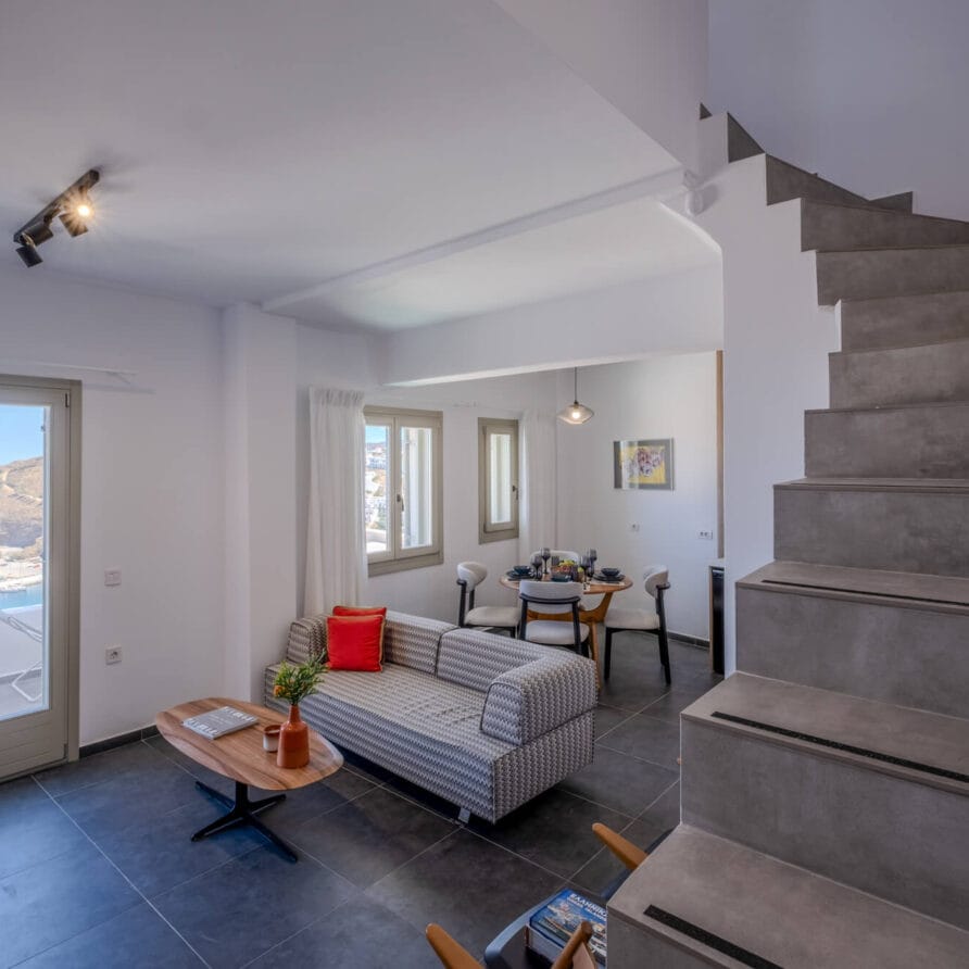Μοντέρνο σαλόνι με γκρι καναπέ, ξύλινο τραπεζάκι σαλονιού, τραπεζαρία και σκάλα. Τα μεγάλα παράθυρα και οι γυάλινες πόρτες προσφέρουν θέα στη θάλασσα και τους λόφους έξω.