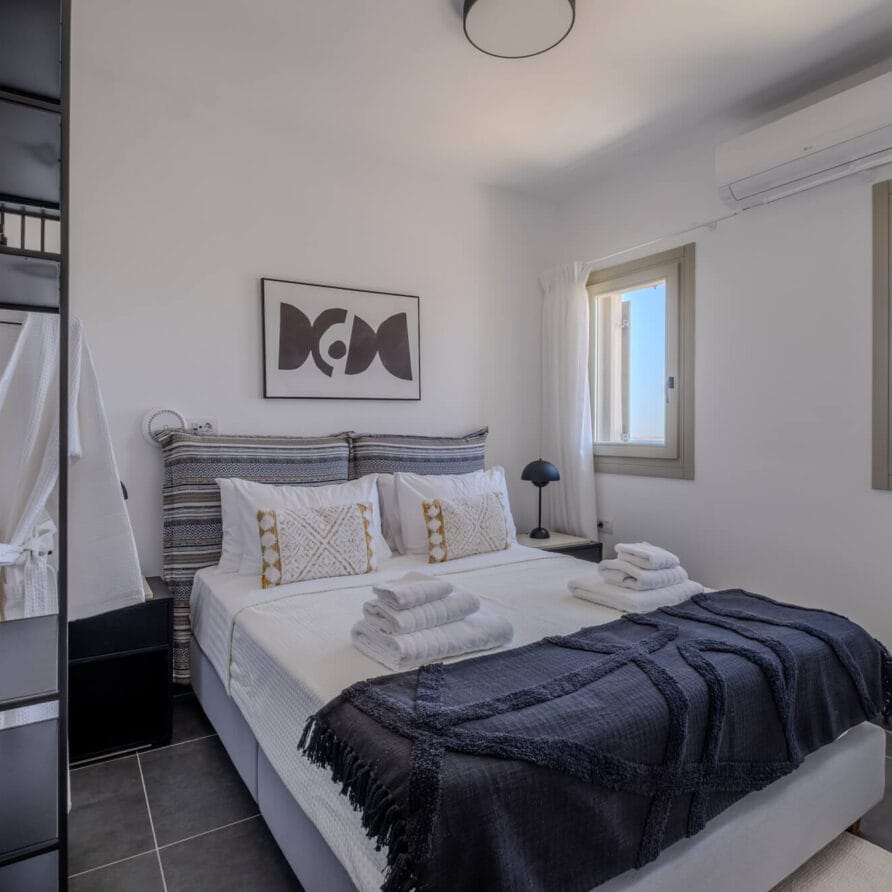 Μοντέρνο υπνοδωμάτιο με διπλό κρεβάτι, πετσέτες διπλωμένες προσεκτικά, δύο παράθυρα, επιτοίχια διακόσμηση πάνω από το κρεβάτι και ένα μαύρο φωτιστικό κομοδίνου σε κομοδίνο. Ρόμπες κρέμονται σε μια κρεμάστρα στα αριστερά.