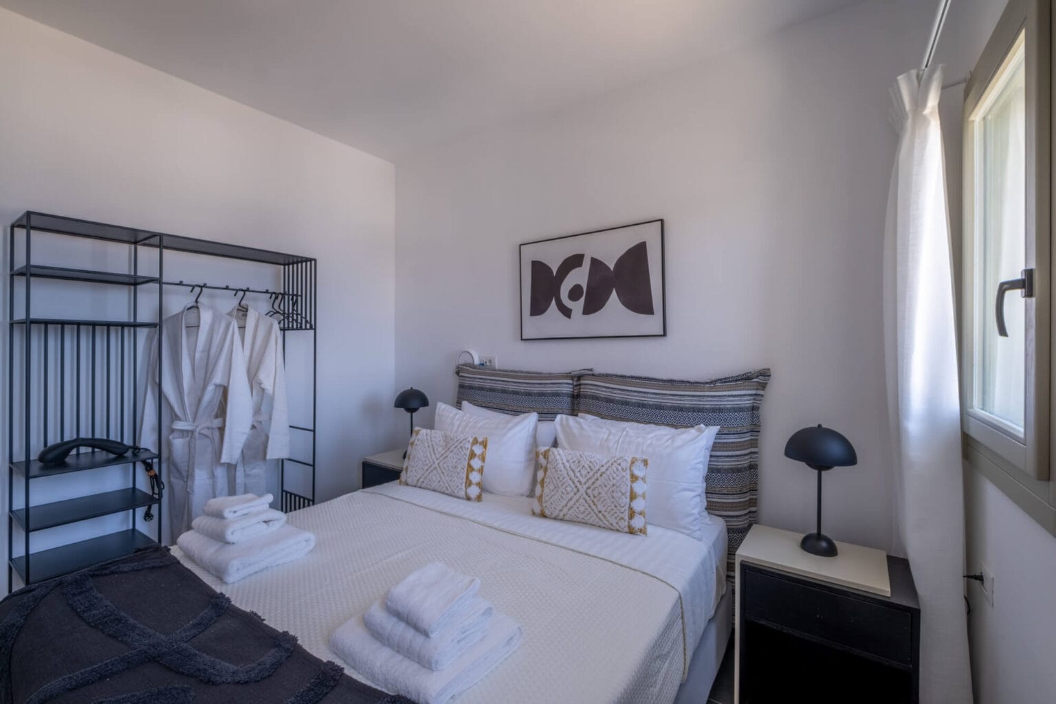 A neatly made double bed with pillows and towels, two bedside tables with lamps, an open wardrobe with robes, and a window with white curtains in a modern bedroom.