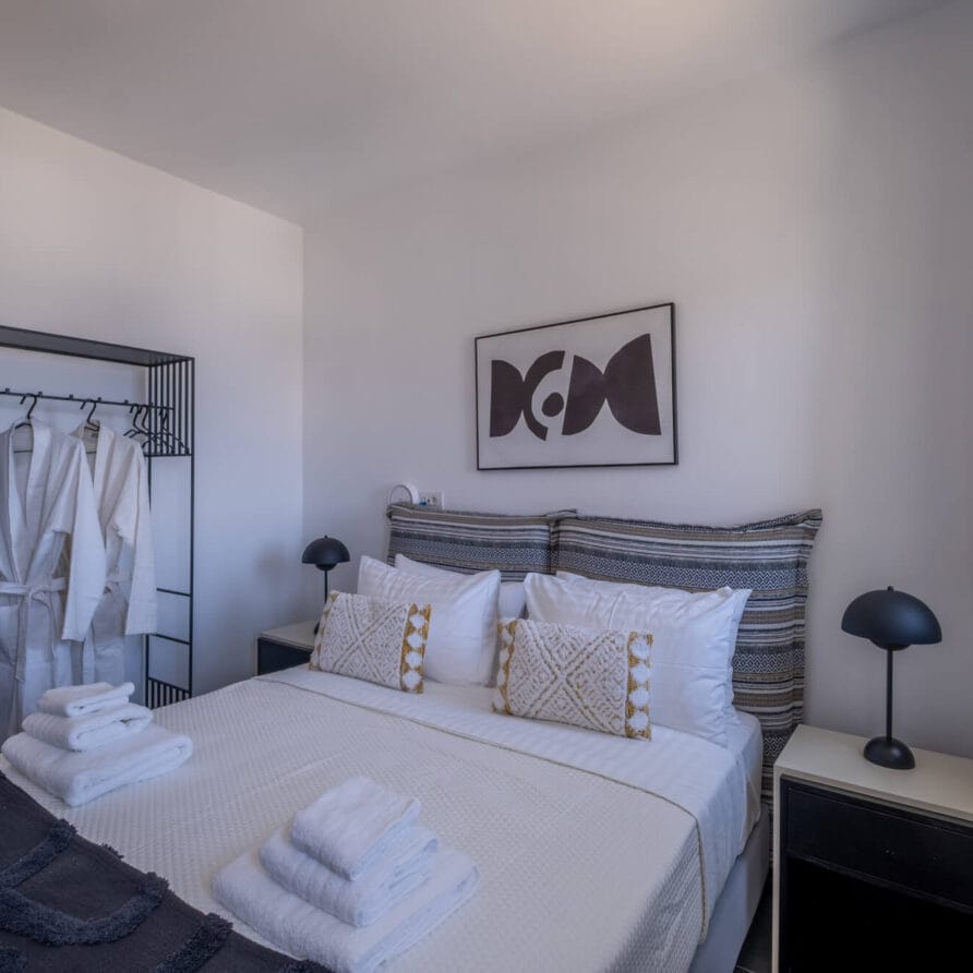 Ένα προσεγμένο κρεβάτι με μαξιλάρια και πετσέτες βρίσκεται σε ένα μικρό, μοντέρνο υπνοδωμάτιο με δύο κομοδίνα, ένα έργο τέχνης τοίχου και μια ανοιχτή ντουλάπα με μπουρνούζια και κρεμάστρες.