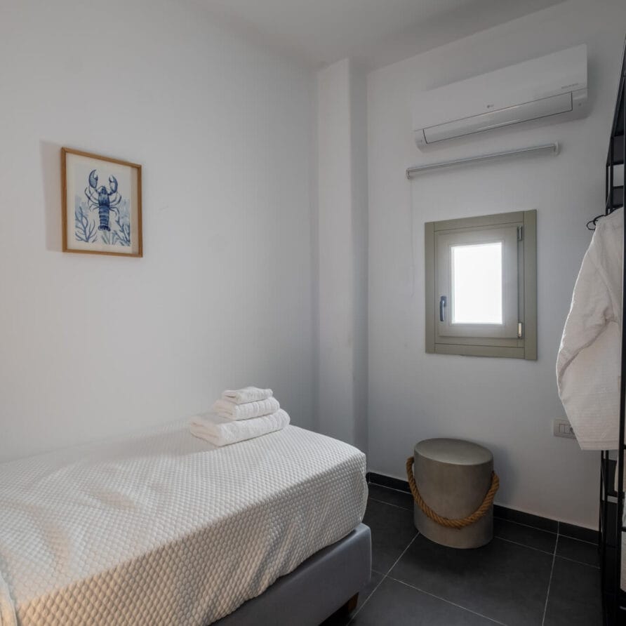 Ένα μικρό, μινιμαλιστικό υπνοδωμάτιο με μονό κρεβάτι, διπλωμένες πετσέτες, έργα τέχνης στον τοίχο, ένα παράθυρο, κλιματιστικό, κρεμαστή λευκή ρόμπα και ένα μικρό στρογγυλό σκαμπό.
