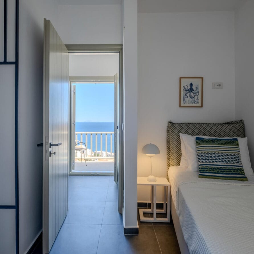 Ένα μικρό υπνοδωμάτιο με μονό κρεβάτι, λευκούς τοίχους, ένα κομοδίνο με φωτιστικό, μια ανοιχτή πόρτα και θέα στη θάλασσα και το μπαλκόνι από την πόρτα.