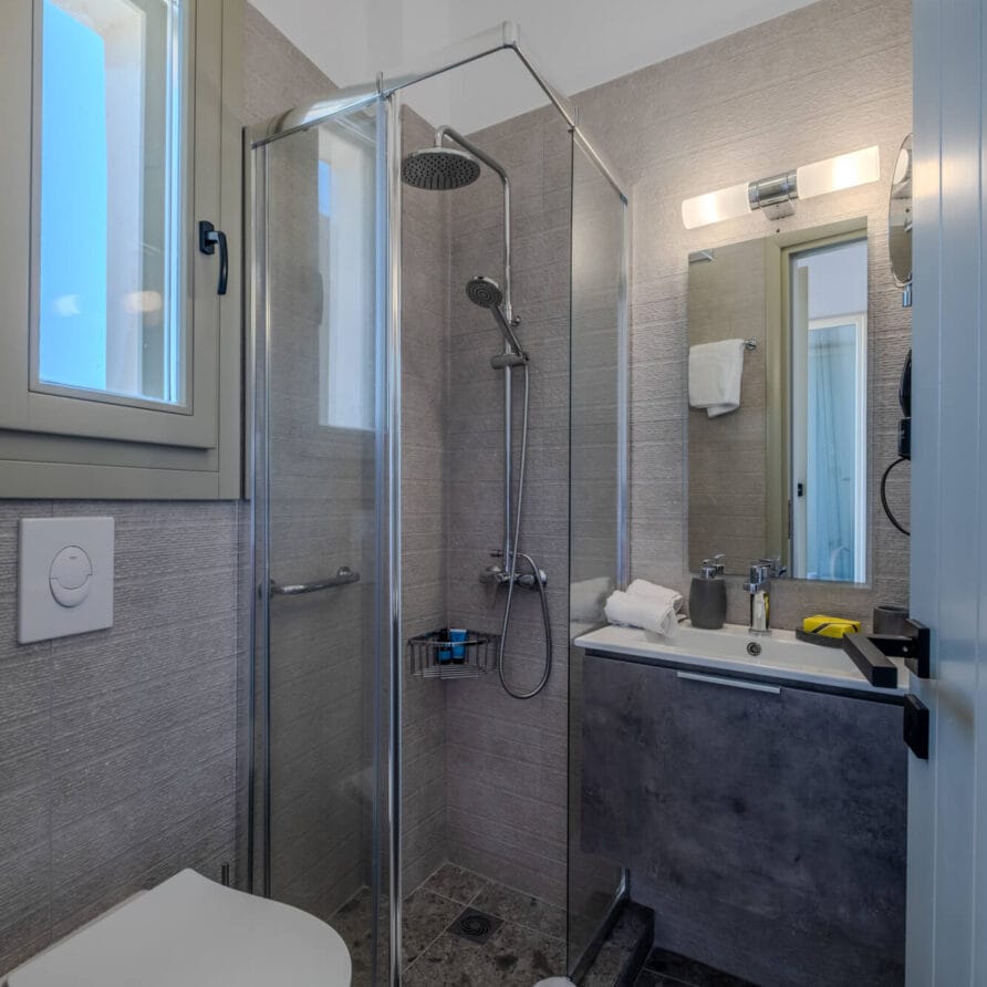 Μοντέρνο μπάνιο με γυάλινη καμπίνα ντους, επιτοίχια τουαλέτα, νιπτήρα με ντουλάπι, καθρέφτη με φως, παράθυρο και πετσέτα στο πάτωμα.