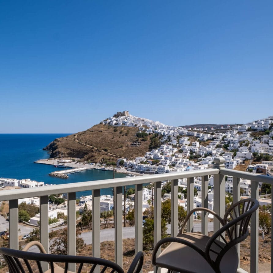 Μπαλκόνι με θέα σε έναν κόλπο, μια πόλη στην πλαγιά του λόφου με λευκά κτίρια και τη γαλάζια θάλασσα κάτω από έναν καθαρό ουρανό. Δύο καρέκλες υπάρχουν στο μπαλκόνι.