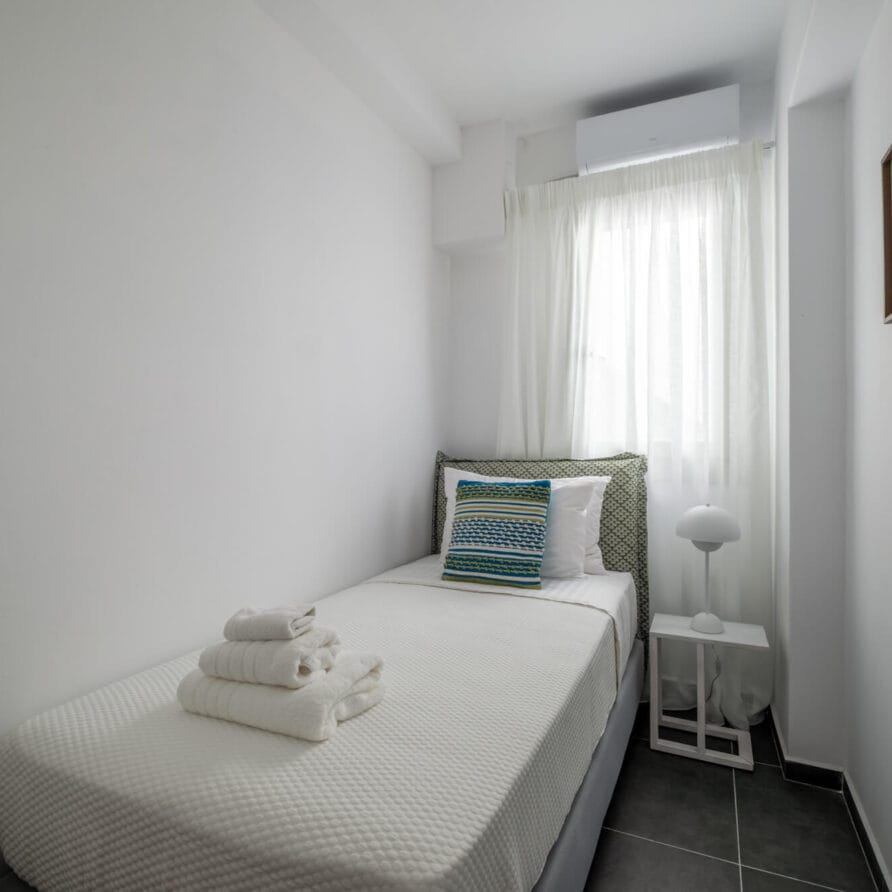 Ένα μικρό, φωτεινό υπνοδωμάτιο με ένα μονό κρεβάτι, διπλωμένες πετσέτες, δύο κορνιζαρισμένες εικόνες στον τοίχο, ένα παράθυρο με διαφανείς κουρτίνες και ένα λευκό κομοδίνο με ένα φωτιστικό.