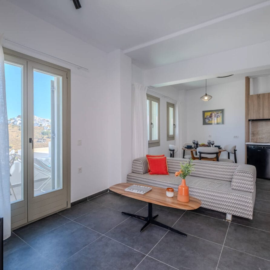 Μοντέρνο σαλόνι με γκρι καναπέ, τραπεζάκι σαλονιού και μικρή κουζίνα. Το φως του ήλιου μπαίνει μέσα από γυάλινες πόρτες και ράφια με πιάτα είναι ορατά στο βάθος.