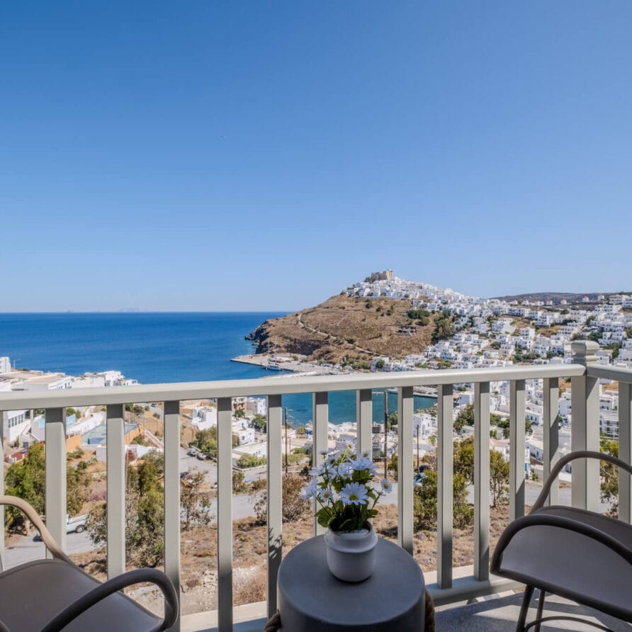 Μπαλκόνι με δύο καρέκλες και ένα μικρό τραπέζι με θέα σε μια παραθαλάσσια πόλη, τη γαλάζια θάλασσα και την πλαγιά ενός λόφου κάτω από έναν καθαρό ουρανό.