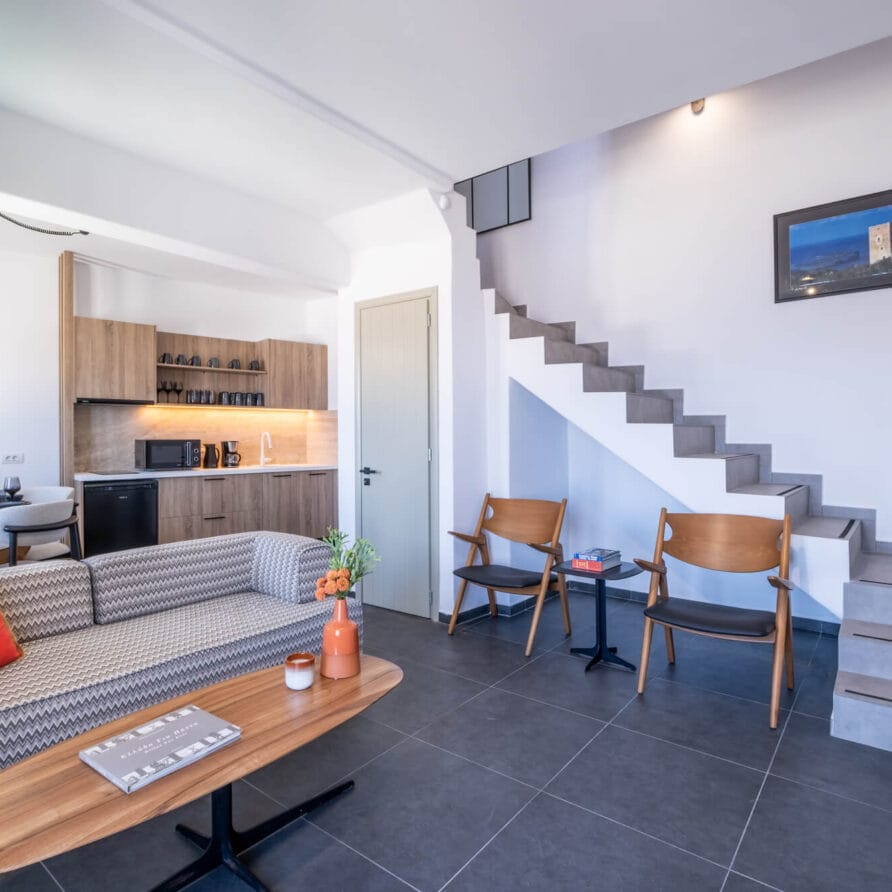 Μοντέρνο σαλόνι με γκρι πλακάκια δαπέδου, καναπέ με σχέδια, ξύλινες καρέκλες, τραπεζάκι σαλονιού, μικρή κουζίνα, τραπεζαρία και σκάλα που οδηγεί στον επάνω όροφο. Η διακόσμηση περιλαμβάνει έργα τέχνης τοίχου και βάζα.