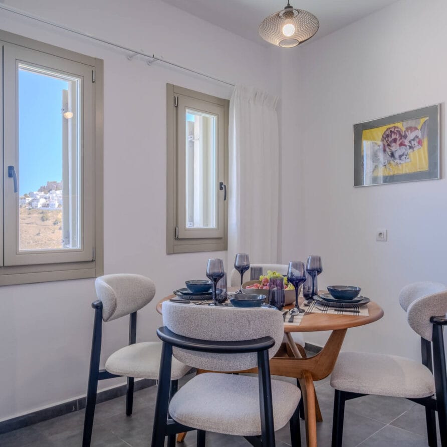 Μια μικρή τραπεζαρία με ένα στρογγυλό ξύλινο τραπέζι για τέσσερα άτομα, περιτριγυρισμένη από ανοιχτόχρωμες γκρι καρέκλες, κοντά σε δύο παράθυρα με ηλιόλουστη θέα προς τα έξω.