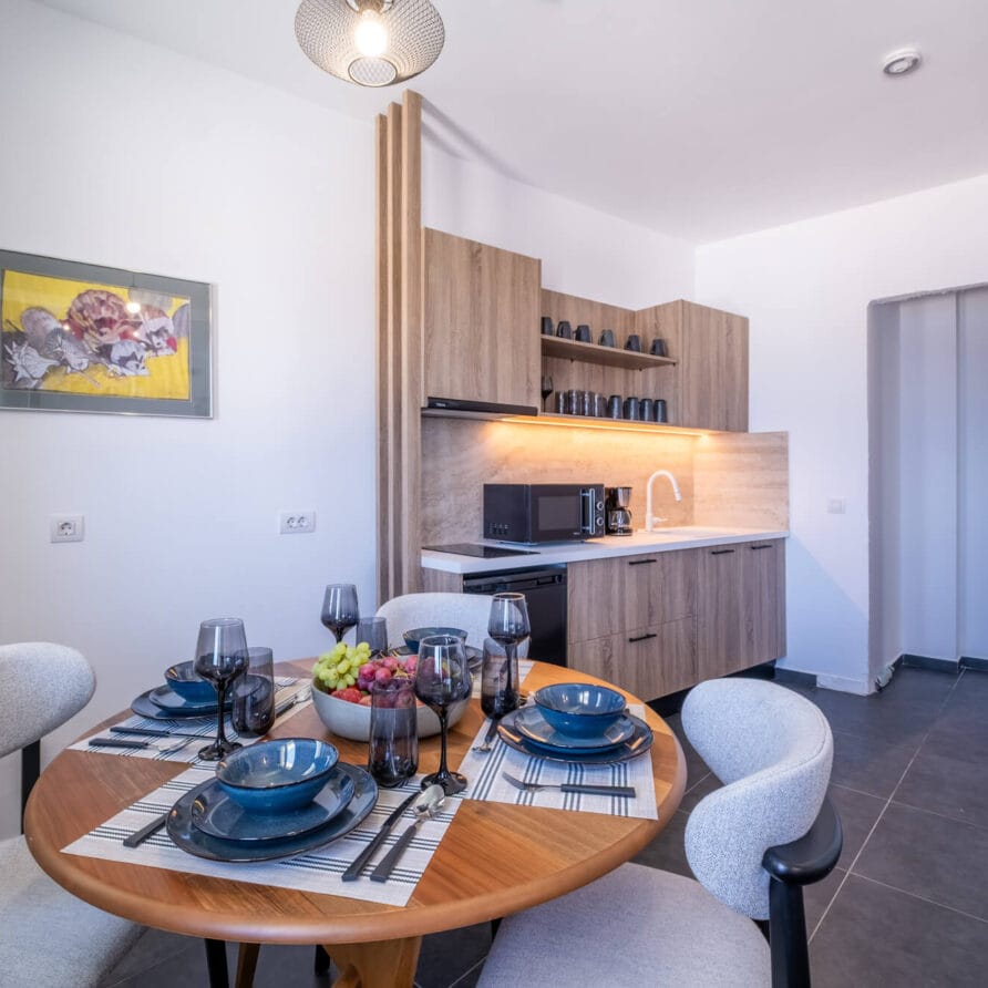 Μια μικρή μοντέρνα κουζίνα με ξύλινα ντουλάπια, ένα στρογγυλό τραπέζι φαγητού για τέσσερα άτομα και ένα κεντρικό μπολ με φρούτα· έργα τέχνης κρέμονται σε έναν λευκό τοίχο.