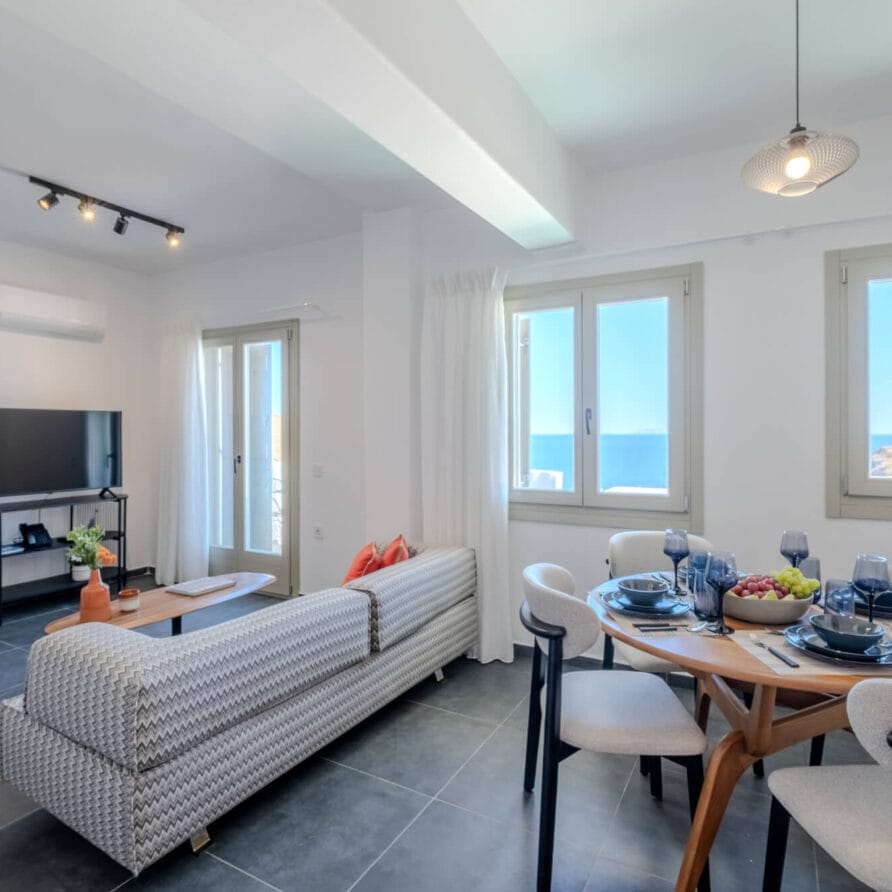 Μοντέρνος χώρος καθιστικού και τραπεζαρίας με καναπέ με σχέδια, στρογγυλό τραπέζι φαγητού για τέσσερα άτομα, τηλεόραση επίπεδης οθόνης και μεγάλα παράθυρα που επιτρέπουν την είσοδο φυσικού φωτός.