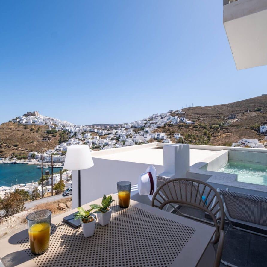 Εξωτερική βεράντα με τραπέζι στρωμένο για δύο άτομα, ποτά και φυτά, με θέα σε μια πισίνα και μια πόλη στην πλαγιά του λόφου με λευκά κτίρια κοντά στη θάλασσα κάτω από έναν καταγάλανο ουρανό.