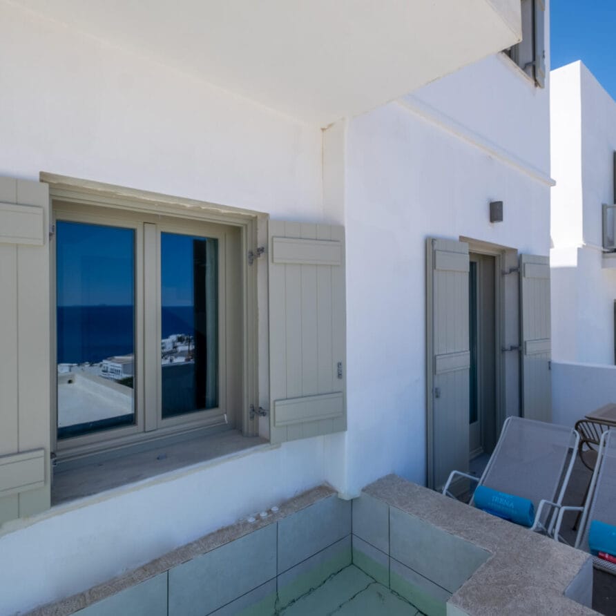 Λευκό εξωτερικό κτίριο με μπεζ παντζούρια, δύο ξαπλώστρες, ένα τραπέζι και μια μικρή πισίνα σε μπαλκόνι με θέα σε μια πλαγιά κάτω από έναν καταγάλανο ουρανό.