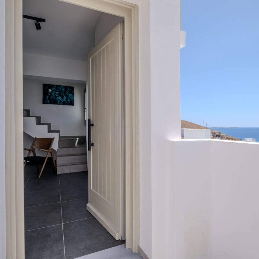 Ανοιχτή πόρτα που οδηγεί σε ένα μοντέρνο εσωτερικό με σκάλες και μια καρέκλα, με θέα σε ένα μπαλκόνι με θέα στη θάλασσα κάτω από έναν καθαρό ουρανό.