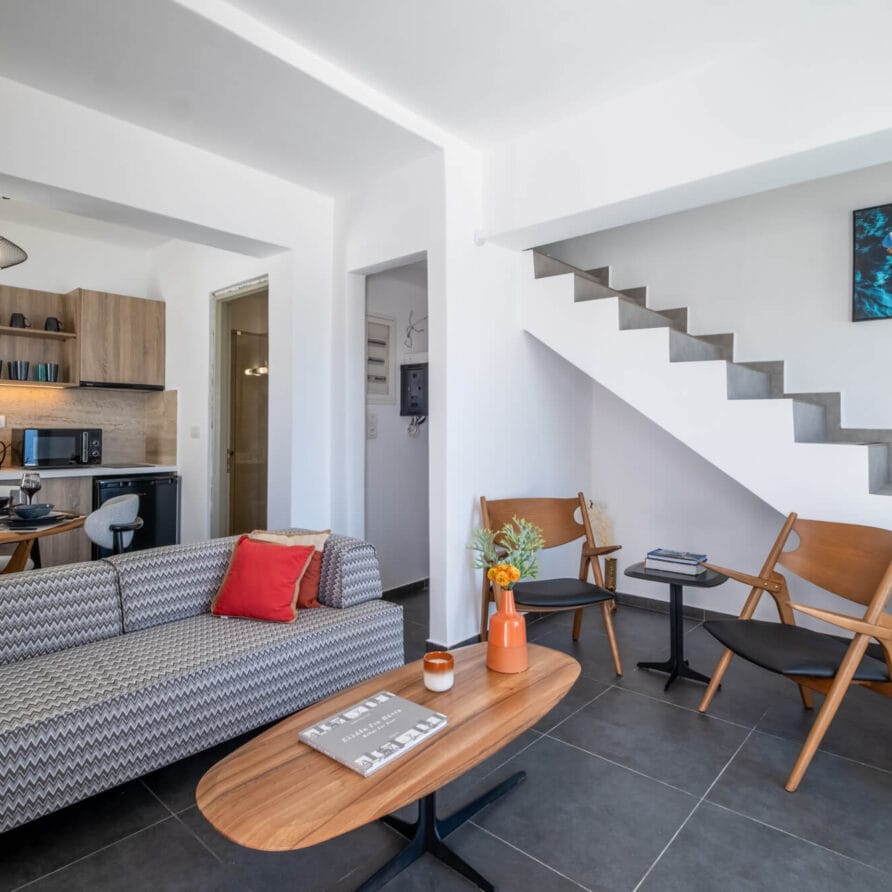 Μοντέρνο σαλόνι με γκρι καναπέ, ξύλινο τραπεζάκι σαλονιού, δύο καρέκλες και ανοιχτή κουζίνα. Η σκάλα οδηγεί προς τα πάνω και έργα τέχνης κρέμονται στον τοίχο.