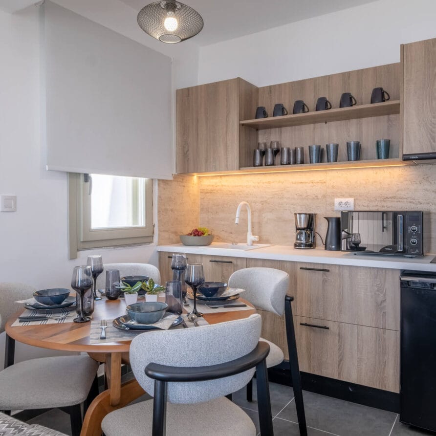 Μοντέρνα κουζίνα και τραπεζαρία με ξύλινα ντουλάπια, στρογγυλό τραπέζι για τέσσερα άτομα, ανοιχτά ράφια με κούπες, συσκευές πάγκου και ένα μικρό ψυγείο κάτω από τον πάγκο.