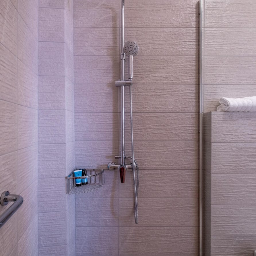 Μοντέρνο ντους με κεφαλή ντους ψιλής βροχής, ντους χειρός, επιτοίχια θήκη με προϊόντα περιποίησης και τοίχους με μπεζ ανάγλυφα πλακάκια.